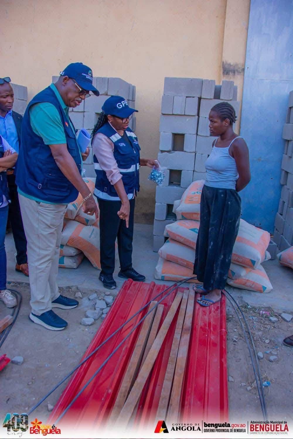 Várias pessoas conversam perto de materiais de construção, incluindo chapas de metal, tábuas de madeira, sacos de cimento e blocos de concreto. Uma mulher se destaca, enquanto outras usam coletes e chapéus com logotipos oficiais.