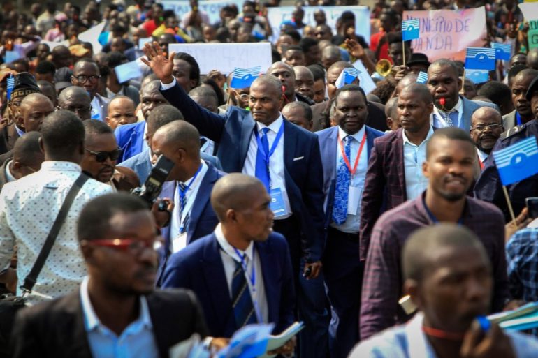 Um grande grupo de pessoas, a maioria homens de terno, marcham juntos segurando pequenas bandeiras azuis e brancas. Um homem no centro acena com a mão e alguns participantes carregam cartazes. A atmosfera parece enérgica e organizada.